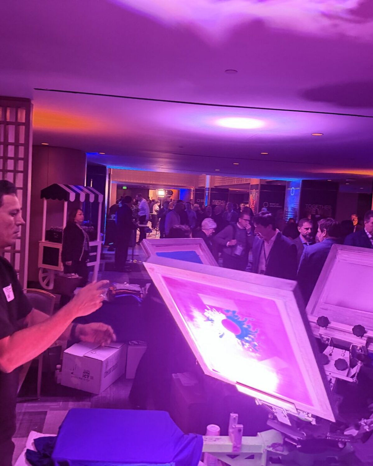 Merch Troop printer working a magenta-lit screen-print press while event guests watch
